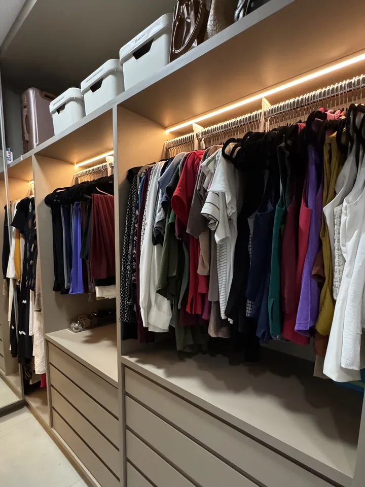 Closet planejado com fita de LED sob as prateleiras, arara de roupas agrupadas por cor, caixas brancas na prateleira de cima e gavetas embutidas embaixo